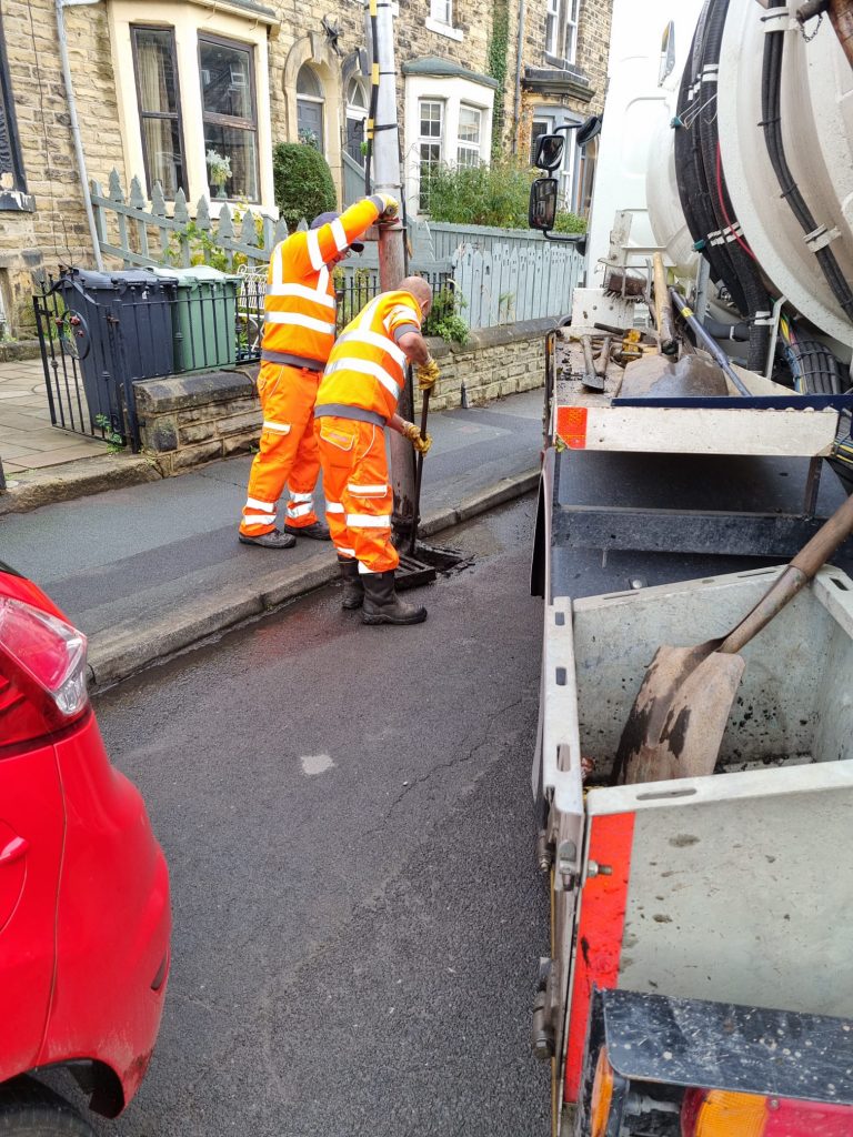 Workers in orange hi-viz overalls operating vehicle-mounted drain clearing equipment, lowering a hose into the opened road drain