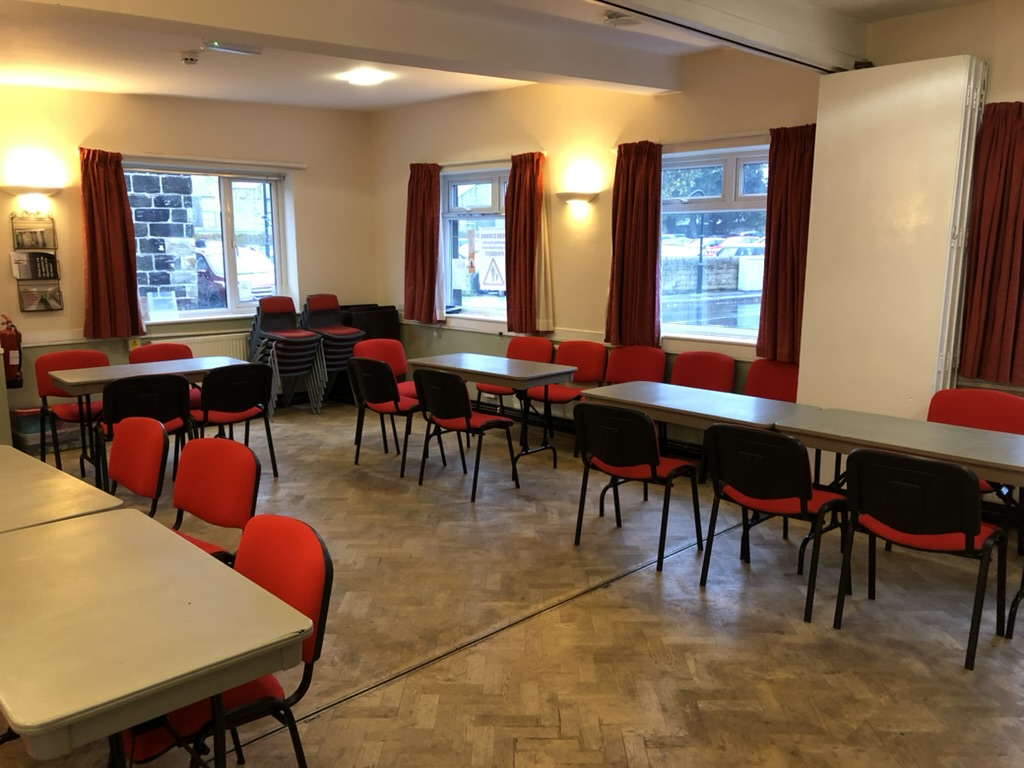 Room with wooden floor showing five plastic folding tables and stackable upholstered chairs