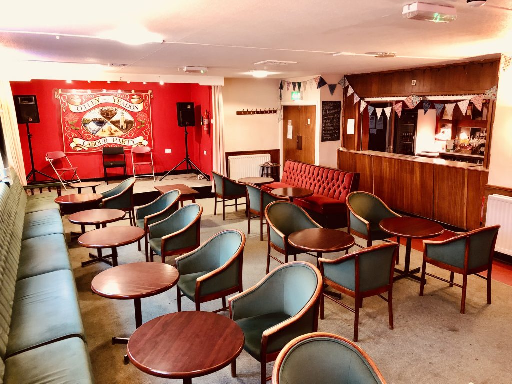 A bright view of the room. The walls a cream/white. Down the window side light green upholstered sofas, another side a small red upholstered sofa. In the middle 9 small round wooden tables, and 12 seats upholstered the same light green. To the right a simple bar with flat wood panels, with bunting over it. At the back an alcove with a small stage with the wall painted red and a painted mural of Otley and Yeadon Labour Party reminiscent of an old marching banner, red and gold, with roses and four images: the Otley clock tower, a field of cut corn, a weir in a river, a printing press. There the two PA speakers and three simple chairs on the stage.