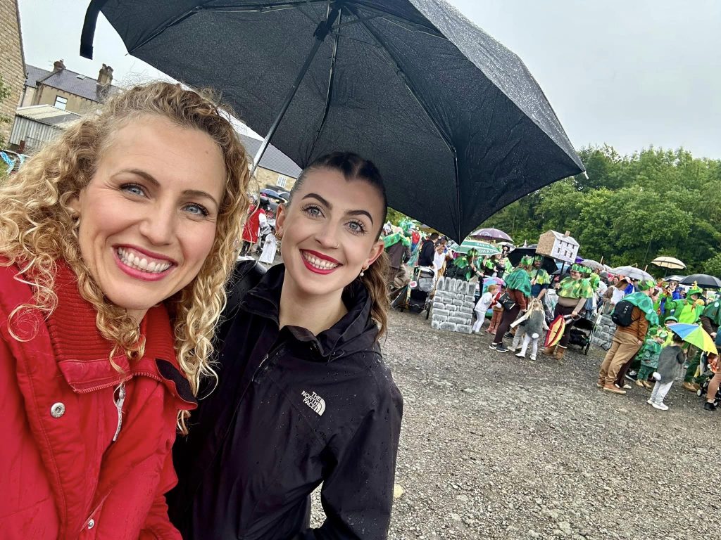 Katie and Sylvia smiling under an umbrella as the carnival gets ready to form up behind them