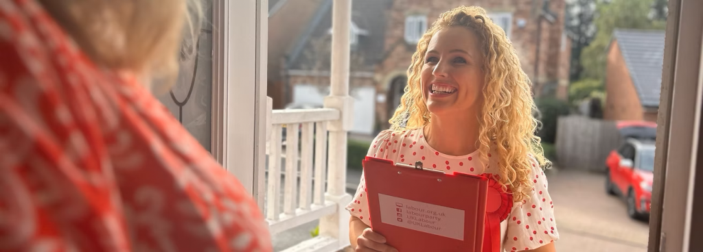 We're looking over someone's shoulder, their front door is open and Katie White is on their doorstep smiling broadly